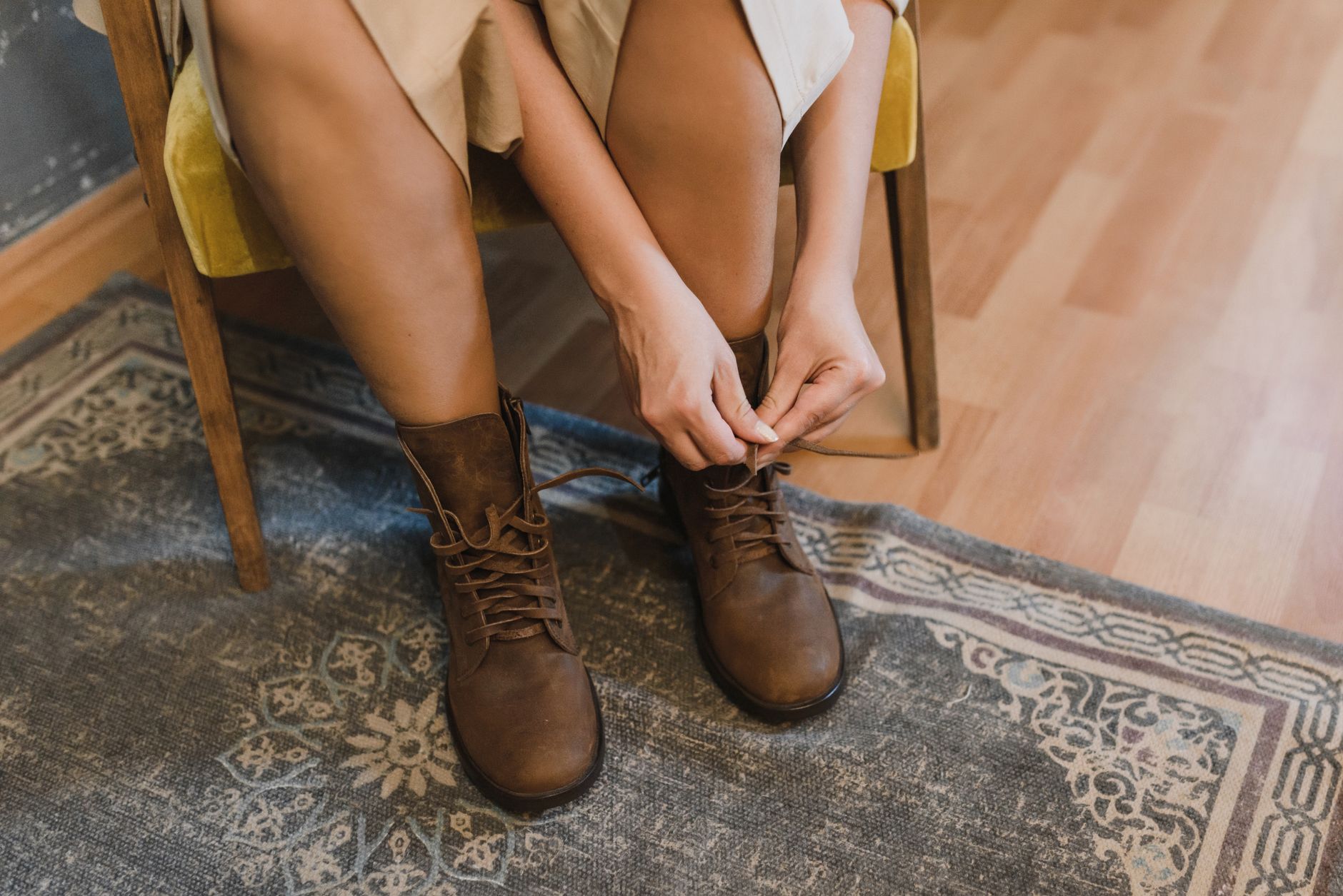 Person sitting on wooden chair lacing up brown leather lace-up boots over an ornate area rug